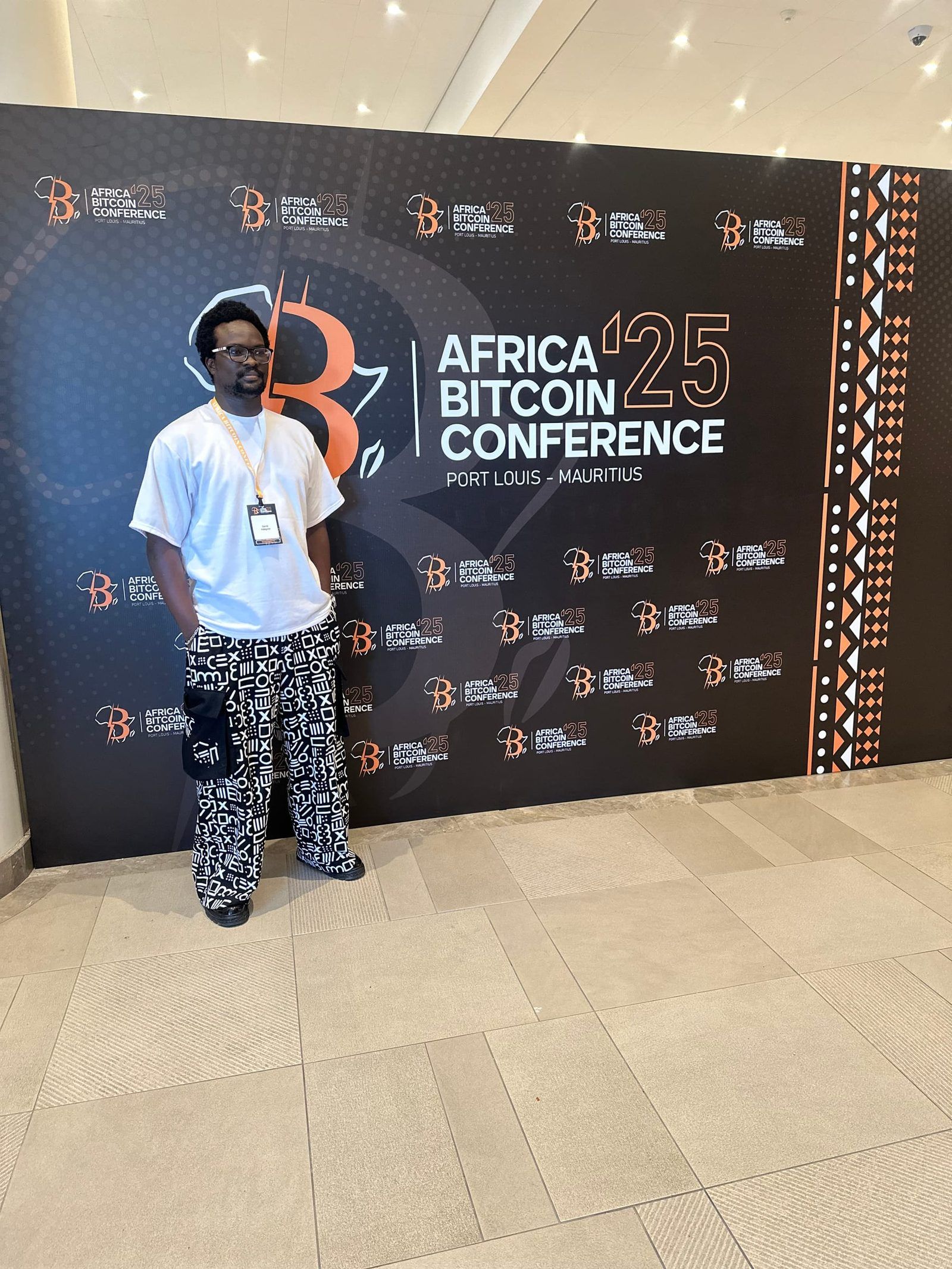 David at the Africa Bitcoin Conference '25 step-and-repeat in Port Louis, attendee lanyard on.