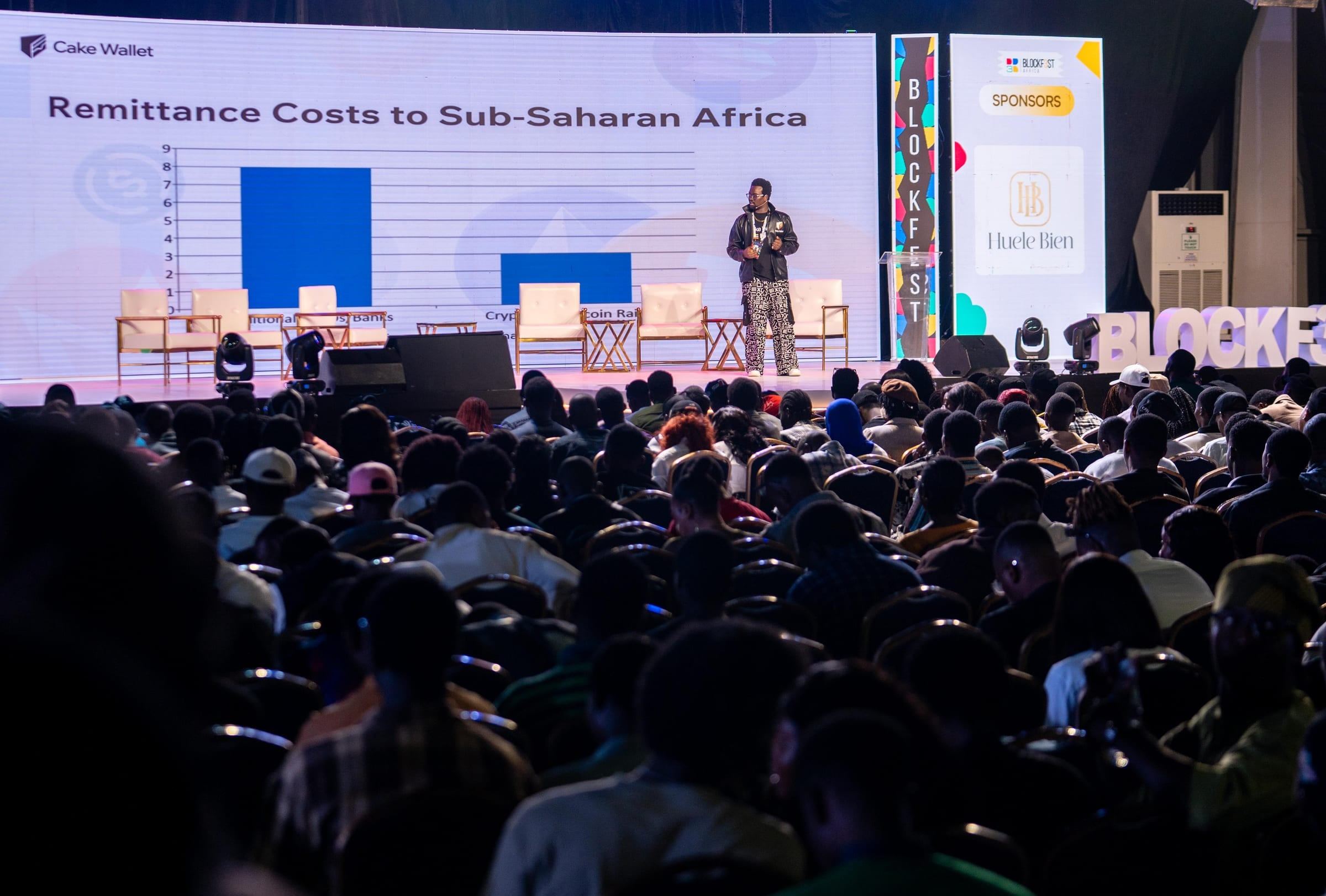 David Adegoke on the main stage at Blockfest Africa 2025, seen past the seated hall, with a slide on remittance costs to Sub-Saharan Africa behind him.