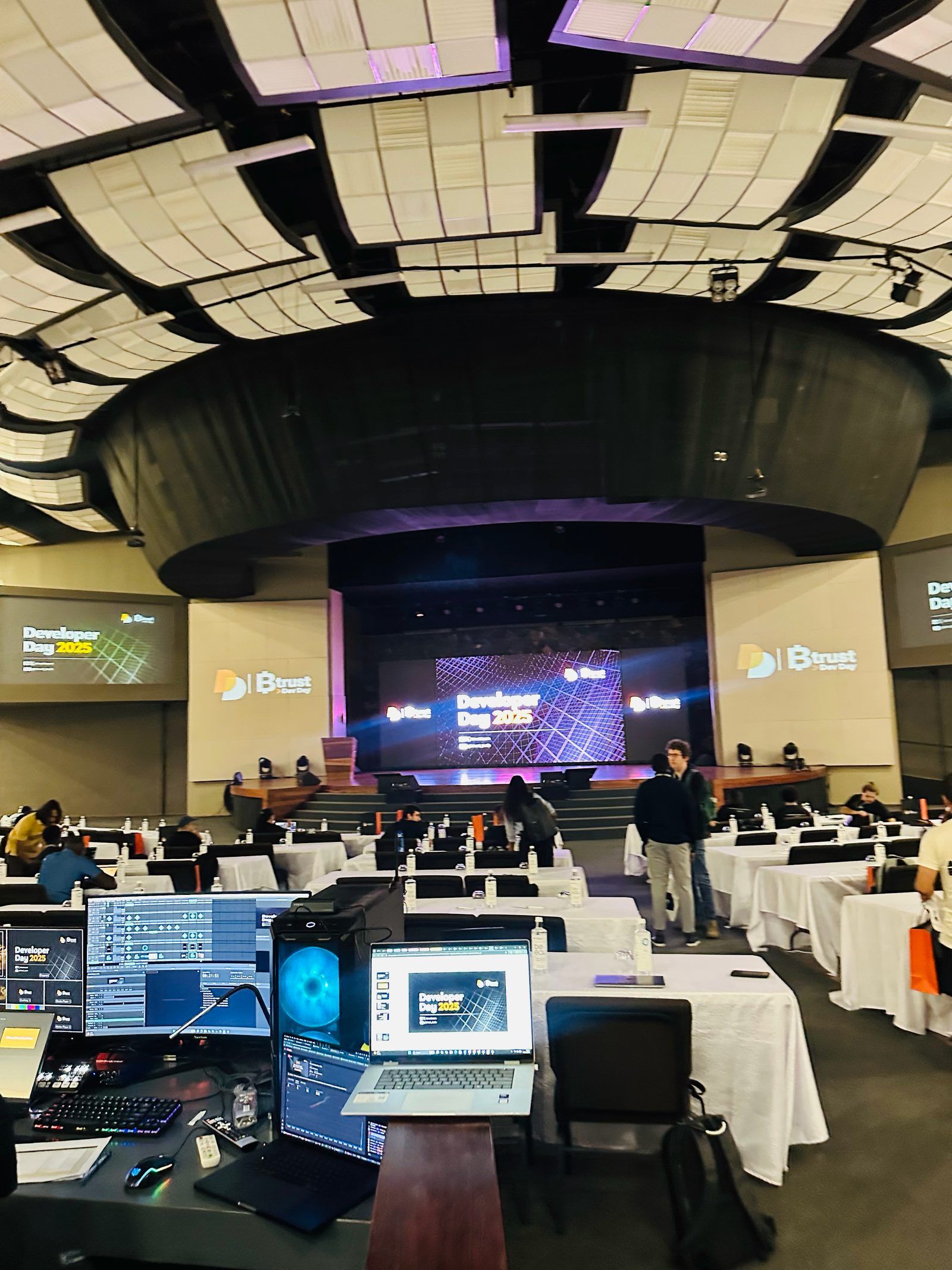 The Btrust Developer Day 2025 hall at the Trianon Convention Centre, Developer Day 2025 on the stage screen and rows of laptops on the dev tables.