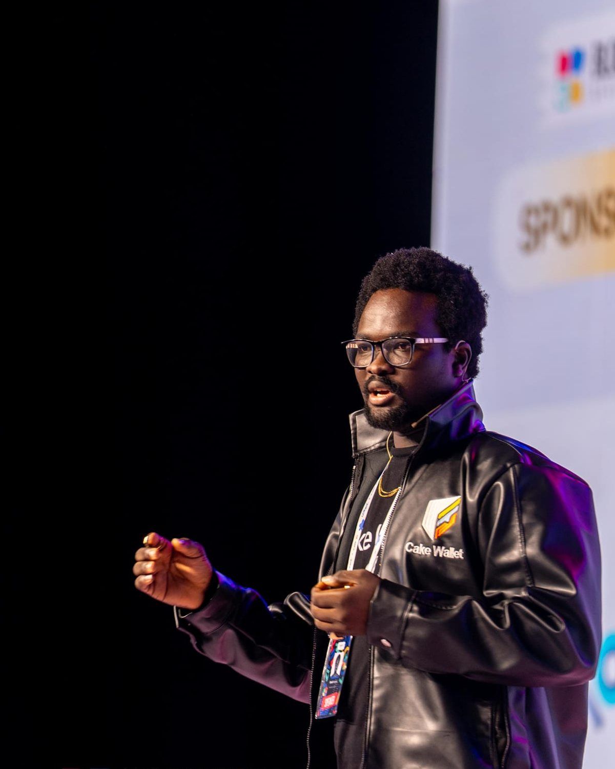 David Adegoke on stage at Blockfest Africa 2025 in Lagos, mid-sentence in a Cake Wallet jacket with a headset mic.